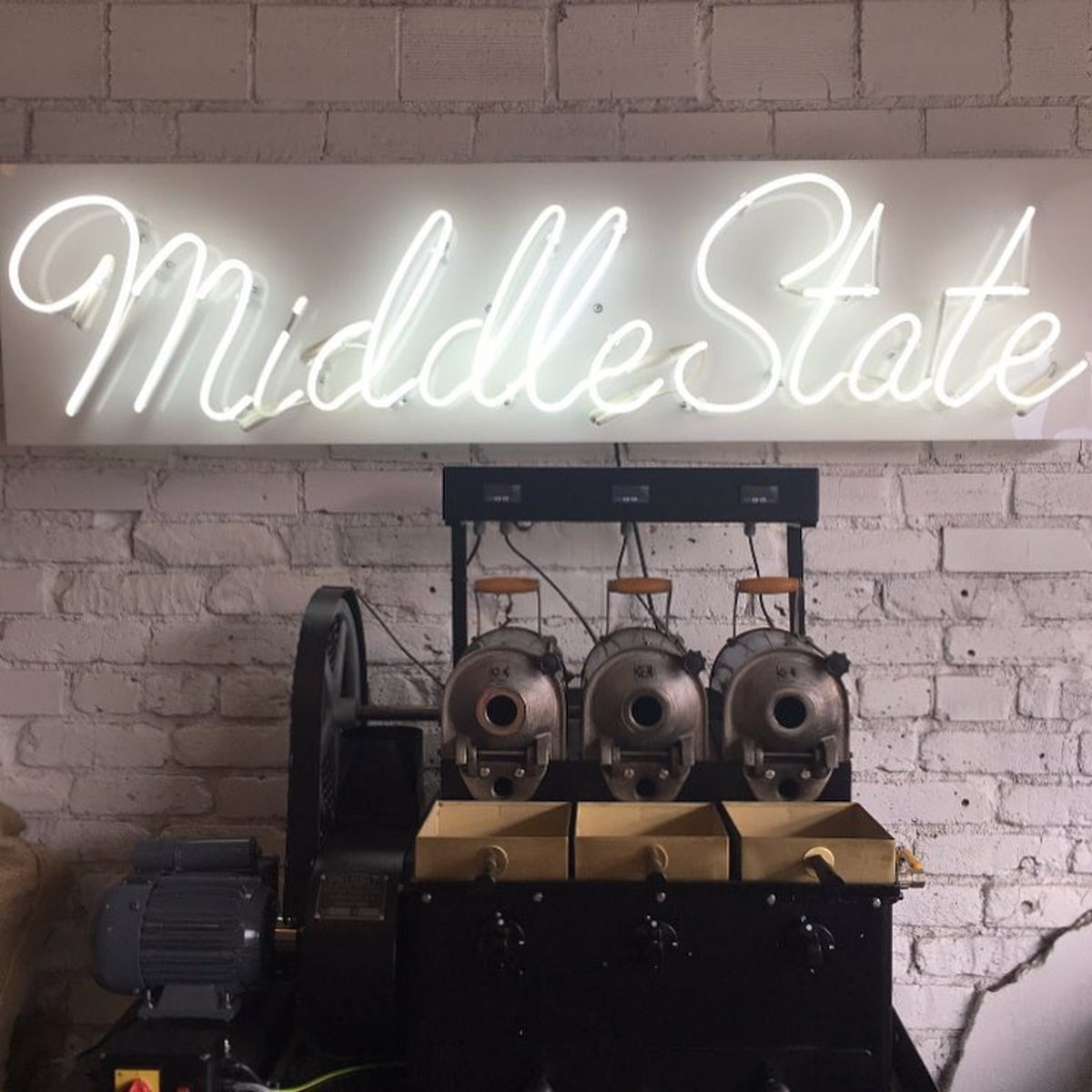 White script neon sign mounted on a brick wall above coffee roasting equipment